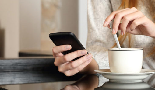 Person rører i kaffe og ser på mobilen. Foto.