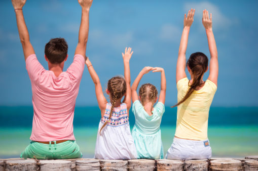 Foto: Småbarnsfamilie sitter på en brygge og ser ut over havet, alle jubler og har hendene over hodet.