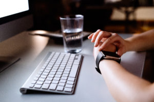 Foto: Venstre arm hviler på pulten og personen ser på klokken. Tastatur og skjerm ved siden av, sen kveld