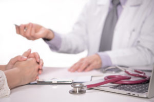 Foto: Legekonsultasjon, stetoskop og journal ligger mellom lege og pasient,. Man ser kun hendene