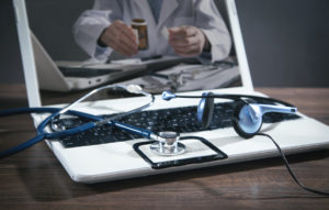 Foto: E-konsultasjon med lege, Laptop med bilde av lege. PÅ tastaturet ligger stetoskop og headset.