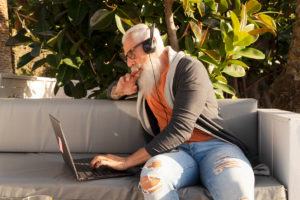 Foro: Eldre mann med headset, jobber avslappet på PCen på en benk i en park eller kafé´. Han er ungdommelig kledd.