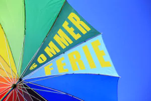 Foto: Fargerik parasoll mot blå himmel. På parasollen står det sommerferie