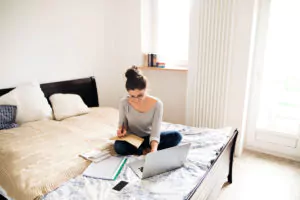 Foto: Dame sitter på sengen og jobber med PC, papirer og telefon